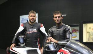 Two men in motorcycle suits standing by a motorcycle holding helmets|